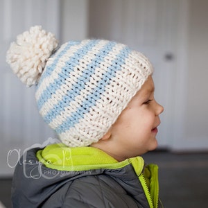 以下が含まれることがあります： 白と青のストライプのニット帽をかぶった子供。帽子の上には白いポンポンがついています。帽子は子供の耳と額にまでかぶっています。子供は緑と灰色のジャケットを着ています。