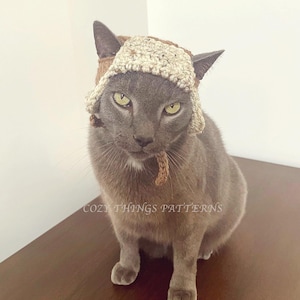 May include: A gray cat wearing a brown and beige crocheted hat with a long brown tassel. The cat is sitting on a wooden surface. The text "Cozy Things Patterns" is visible on the surface.