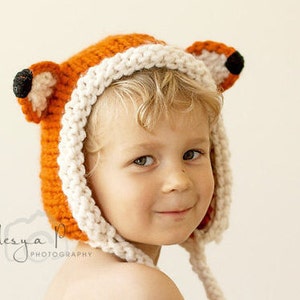 May include: A child wearing a knitted fox hat with orange and white fur. The hat has large, floppy ears and a white pom-pom on top.