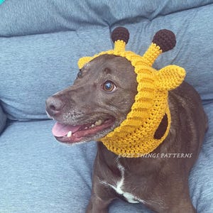 Könnte beinhalten: Ein brauner Hund trägt ein gelbes, gehäkeltes Giraffenkostüm mit braunen Ohren. Das Kostüm hat einen schwarzen Fleck an der Seite. Der Hund sitzt auf einem grauen Sofa. Der Text "Cozy Things Patterns" ist auf dem Kostüm sichtbar.