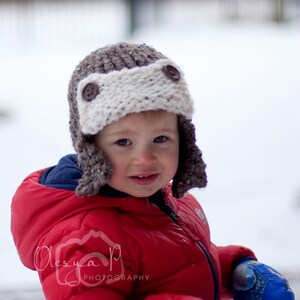 May include: A young child wearing a brown and white knitted aviator hat with brown buttons and a red padded jacket. The child is standing in the snow.