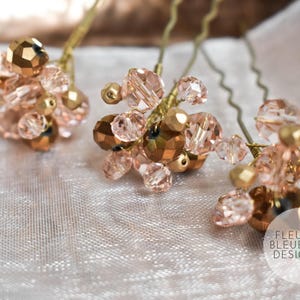 Peut inclure: Trois &eacute;pingles &agrave; cheveux orn&eacute;es de grappes de perles facett&eacute;es rose clair et bronze m&eacute;tallis&eacute; fix&eacute;es sur des tiges en fil dor&eacute;. Chaque &eacute;pingle poss&egrave;de une base m&eacute;tallique ondul&eacute;e. Un logo circulaire avec le texte FLEUR BLEUE DESIGN.com est visible.