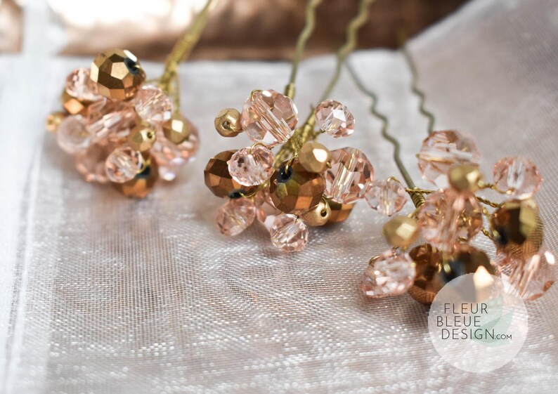 Peut inclure: Trois &eacute;pingles &agrave; cheveux orn&eacute;es de grappes de perles facett&eacute;es rose clair et bronze m&eacute;tallis&eacute; fix&eacute;es sur des tiges en fil dor&eacute;. Chaque &eacute;pingle poss&egrave;de une base m&eacute;tallique ondul&eacute;e. Un logo circulaire avec le texte FLEUR BLEUE DESIGN.com est visible.