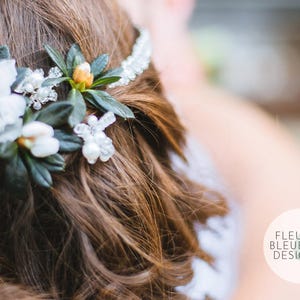 Peut inclure: Un accessoire de coiffure compos&eacute; de fleurs blanches, de feuilles vertes et de perles de cristal transparentes sur un bandeau. L'accessoire est plac&eacute; dans des cheveux bruns. Un logo circulaire affiche FLEUR BLEUE DESIGN.com et une feuille.