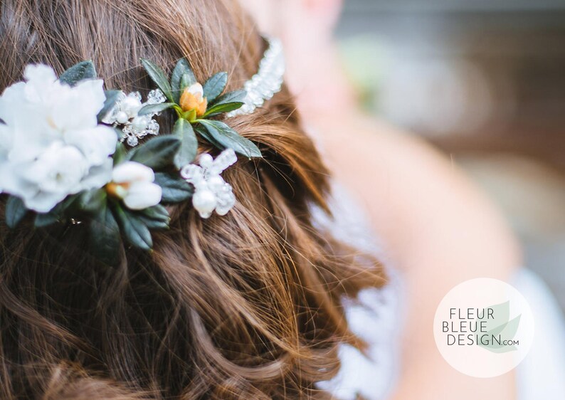 Peut inclure: Un accessoire de coiffure compos&eacute; de fleurs blanches, de feuilles vertes et de perles de cristal transparentes sur un bandeau. L'accessoire est plac&eacute; dans des cheveux bruns. Un logo circulaire affiche FLEUR BLEUE DESIGN.com et une feuille.