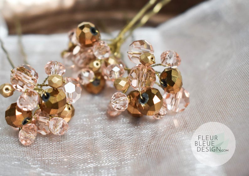 May include: Three hair pins with clusters of rose-gold and gold beads. The beads are faceted and arranged on thin, gold wires. The background is a soft, white fabric. The Fleur Bleue Design logo is in the bottom right corner.