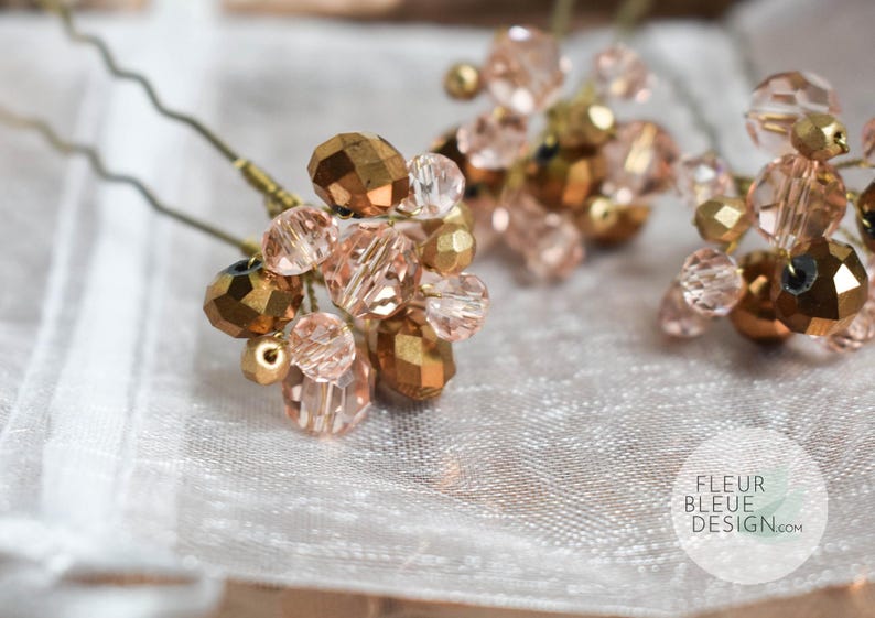 May include: Close-up of several hair pins featuring clusters of faceted, gold and blush-coloured beads. The beads are arranged on thin, wavy, metal pins. The pins rest on a white, textured surface. The Fleur Bleue Design logo is visible.