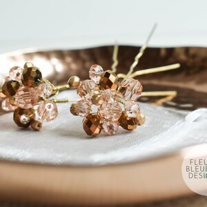 May include: Two hair pins featuring clusters of gold and peach-toned faceted beads. The beads are arranged on gold-coloured wire stems. The pins rest on a white fabric inside a copper-coloured dish.