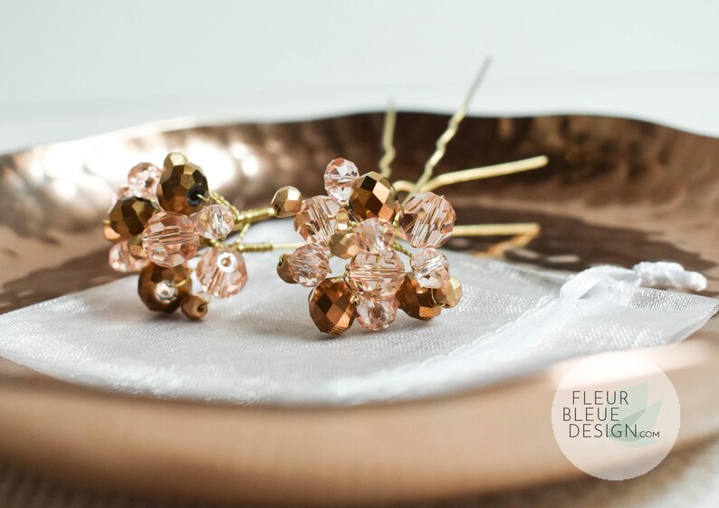 May include: Two hair pins featuring clusters of gold and peach-toned faceted beads. The beads are arranged on gold-coloured wire stems. The pins rest on a white fabric inside a copper-coloured dish.