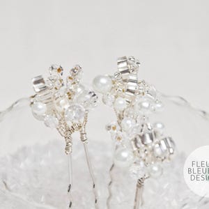 Peut inclure: Deux &eacute;pingles &agrave; cheveux argent&eacute;es orn&eacute;es de grappes de perles blanches artificielles et de cristaux transparents facett&eacute;s fix&eacute;s sur des tiges m&eacute;talliques. Un logo circulaire avec le texte FLEUR BLEUE DESIGN.com figure en bas &agrave; droite.