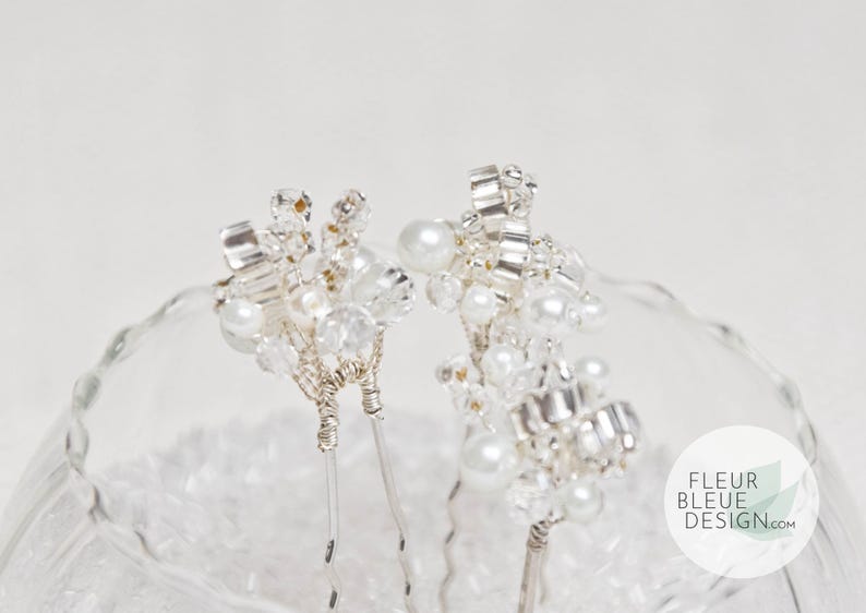 Peut inclure: Deux &eacute;pingles &agrave; cheveux argent&eacute;es orn&eacute;es de grappes de perles blanches artificielles et de cristaux transparents facett&eacute;s fix&eacute;s sur des tiges m&eacute;talliques. Un logo circulaire avec le texte FLEUR BLEUE DESIGN.com figure en bas &agrave; droite.