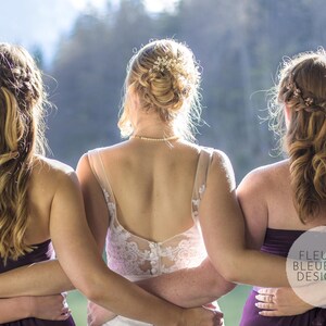 May include: Three women stand together, arms around each other, with their backs to the camera. The woman in the center wears a white lace wedding dress. The other two wear dark purple strapless dresses. Their hair is styled with decorative accents.