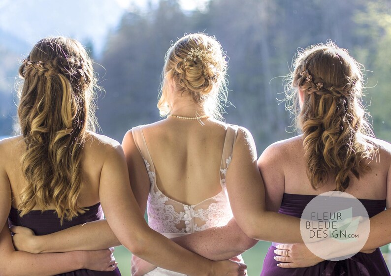 May include: Three women stand together, arms around each other, with their backs to the camera. The woman in the center wears a white lace wedding dress. The other two wear dark purple strapless dresses. Their hair is styled with decorative accents.