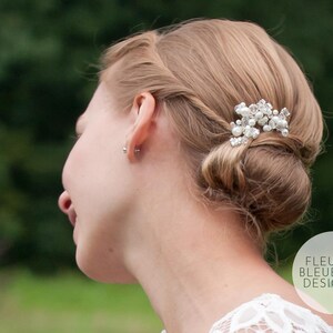 Peut inclure: Un accessoire de coiffure compos&eacute; d'un groupe de perles blanches et de cristaux transparents &agrave; facettes sur une base argent&eacute;e, fix&eacute; dans un chignon blond. Le texte FLEUR BLEUE DESIGN.com est visible dans le coin inf&eacute;rieur droit de l'image.