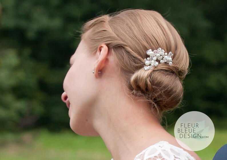 Peut inclure: Un accessoire de coiffure compos&eacute; d'un groupe de perles blanches et de cristaux transparents &agrave; facettes sur une base argent&eacute;e, fix&eacute; dans un chignon blond. Le texte FLEUR BLEUE DESIGN.com est visible dans le coin inf&eacute;rieur droit de l'image.
