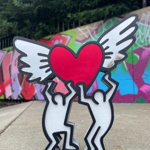May include: A sculpture of a red heart with white wings, supported by two stylized white figures with black outlines. The figures stand on a concrete surface, with graffiti art and trees in the background.