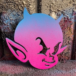May include: A devil-shaped wall art piece with a gradient of blue to pink. The artwork features pointed horns, a smiling face, and pointed ears. The background is a textured, metallic surface with hints of other colours.