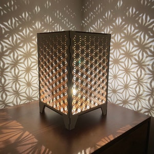 Peut inclure: Une lampe en bois avec un motif géométrique découpé. La lampe est allumée et projette une ombre sur la surface sur laquelle elle est posée.