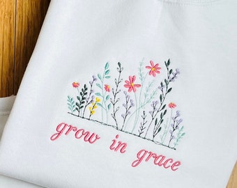 May include: Two white sweatshirts with embroidered floral designs. The designs feature pink, red, and purple flowers with green leaves and stems. The phrase "grow in grace" is embroidered in pink script below the flowers.