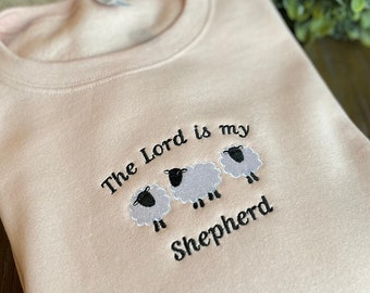 May include: A light pink sweatshirt with the text "The Lord is my Shepherd" embroidered on the front. Three embroidered sheep are below the text.