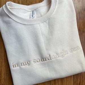 May include: A cream-colored sweatshirt with the text "in my sourdough era" embroidered in a light brown color.