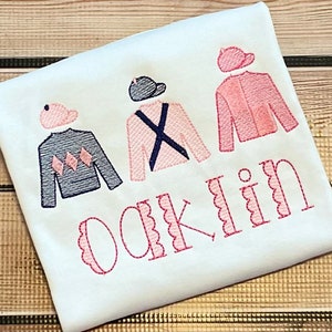 May include: A white t-shirt with three embroidered jockey silks in pink, blue and grey. The silks have a jockey cap on top. The name "Oaklin" is embroidered in pink below the silks.