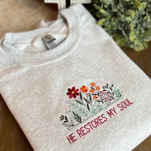 May include: Gray sweatshirt with embroidered flowers and the text "HE RESTORES MY SOUL".