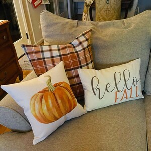 May include: Three throw pillows on a sofa. One pillow has a plaid pattern in brown, white, and orange. Another pillow has a watercolour painting of a pumpkin. The third pillow is white with the text "hello fall" in brown and orange.