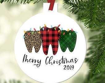 May include: White round Christmas ornament with three teeth decorated in leopard print, red and black plaid, and green plaid. The teeth have string lights around them and the ornament says "Merry Christmas 2019".
