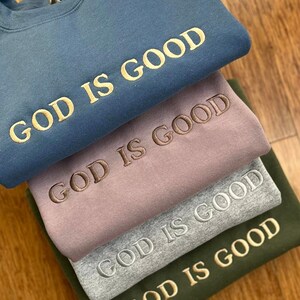 May include: A stack of four sweatshirts in different colours, each with the phrase "God is Good" embroidered in a different coloured thread. The sweatshirts are folded on top of each other, with the blue sweatshirt on top and the green sweatshirt on the bottom.