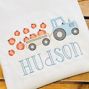 May include: A blue tractor pulls a wagon full of orange pumpkins. The tractor has a white cab and black wheels. The wagon is beige with black wheels. The pumpkins are scattered around the wagon. The name "Hudson" is embroidered in blue thread below the tractor.