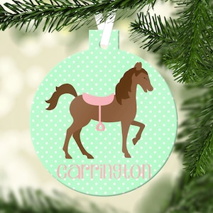 May include: A green and white polka dot Christmas bauble with a brown horse and a pink saddle. The bauble has the name "Carrington" written on it.