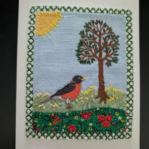 May include: Embroidered greeting card featuring a robin, tree, and sun. The bird has a red breast and black head. The tree has green leaves and a brown trunk. The sun is yellow. The card has a green border and a blue sky background.