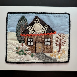 May include: A textile art piece featuring a brown house with a dark roof, white lace accents, and a red trim. The scene includes a light blue sky, a white snowy ground, a green tree, a bare brown tree, and a gray path, all bordered by black stitching.