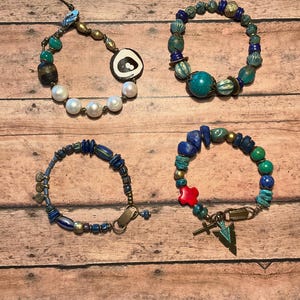 May include: Four beaded bracelets with various designs and colors are displayed on a wooden surface. The bracelets feature a mix of beads, including pearls, turquoise, and blue stones. One bracelet has a large white and black circular bead.