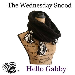 Pode incluir: Cachecol preto de crochet infinito com borlas brancas e pretas. O cachecol é exibido em um manequim com um fundo de tecido de juta. O texto "The Wednesday Snood" e "Hello Gabby" é visível na imagem.