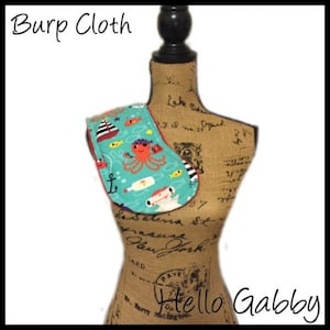 May include: Burp cloth with a nautical theme. The cloth is turquoise with a red border and features an octopus, a ship, a bottle, an anchor, and fish. The text "Hello Gabby" is visible in the bottom right corner.