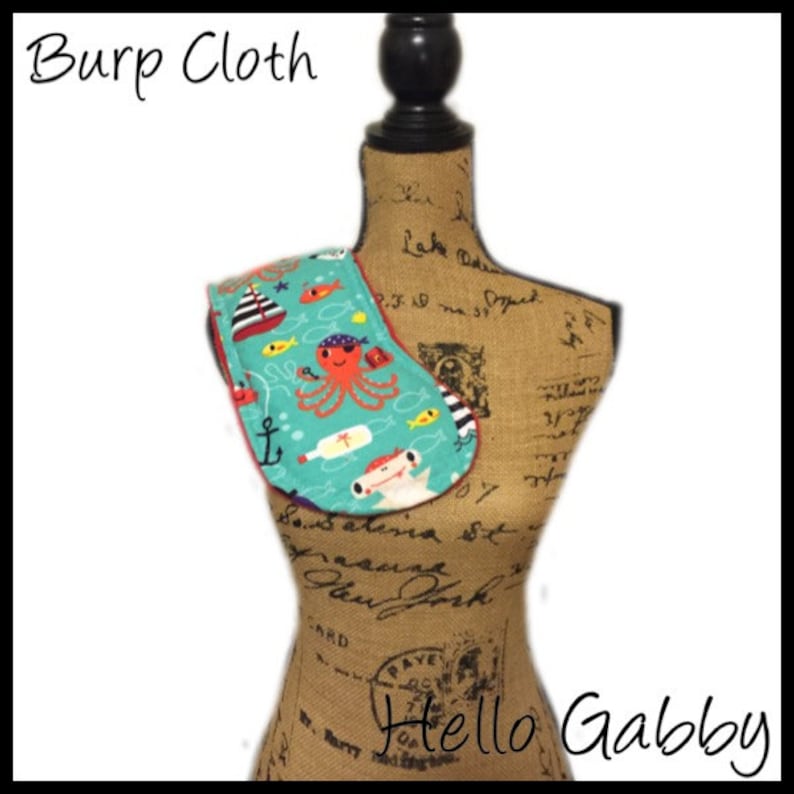 May include: Burp cloth with a nautical theme. The cloth is turquoise with a red border and features an octopus, a ship, a bottle, an anchor, and fish. The text "Hello Gabby" is visible in the bottom right corner.