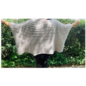K&ouml;nnte beinhalten: Ein beigefarbener und grau gestreifter Poncho mit weiten &Auml;rmeln. Das offene Kleidungsst&uuml;ck besteht aus einem leichten, gestrickten Material. Der Poncho wird mit ausgestreckten Armen pr&auml;sentiert, um das Design und den Fall des Stoffes zu zeigen.