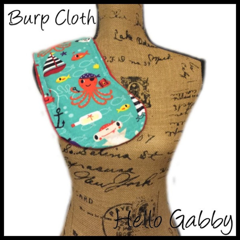 May include: A turquoise and red burp cloth with a nautical theme. The cloth features an octopus, a fish, an anchor, and a bottle with a cross on it. The text "Burp Cloth" is written in black at the top of the image. The text "Hello Gabby" is written in black at the bottom of the image.