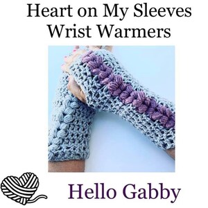 May include: Crocheted wrist warmers in gray with a row of purple flower-like accents. The text "Heart on My Sleeves Wrist Warmers" is at the top, and "Hello Gabby" is at the bottom. The warmers are handmade and designed to keep hands warm.
