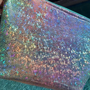 May include: A rectangular iridescent pouch with a textured surface. The pouch has a teal zipper and a shimmering, multi-colored finish, reflecting hues of pink, blue, and green. The overall effect is a sparkly, eye-catching accessory.