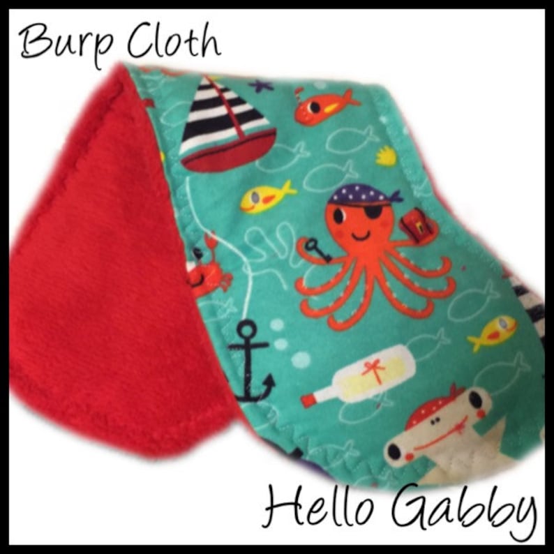 May include: A red and turquoise burp cloth with a nautical theme. The cloth features an octopus with an eye patch, a sailboat, a bottle, an anchor, and fish. The text "Hello Gabby" is printed on the bottom of the cloth.