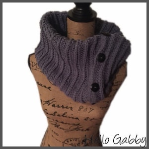 May include: A knitted cowl in a muted purple hue, featuring a ribbed texture and two large black buttons. The cowl is displayed on a mannequin, showcasing its design and fit. The text "Hello Gabby" is visible in the bottom right corner.