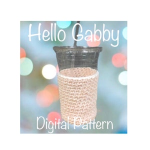 May include: A clear plastic cup with a straw is encased in a beige crocheted cozy. The text "Hello Gabby" is at the top, and "Digital Pattern" is at the bottom. The background has blurred bokeh lights.