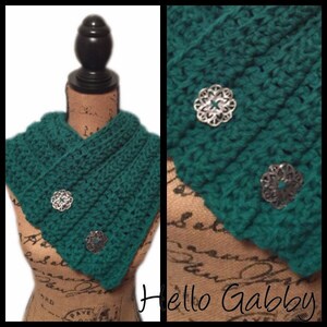 May include: A teal green crocheted cowl with three silver decorative accents. The cowl is displayed on a mannequin.