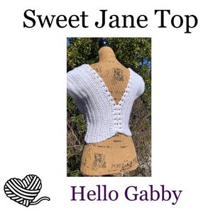 May include: White crochet top with a V-neck and short sleeves. The top is crocheted in a simple stitch and has a scalloped edge. The top is displayed on a mannequin against a blurred background of green trees. The text "Sweet Jane Top" is displayed above the image and "Hello Gabby" is displayed below the image.