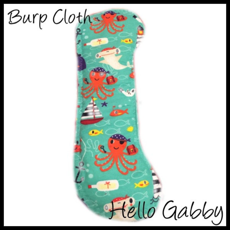 May include: A turquoise burp cloth with a nautical theme featuring a repeating pattern of red octopus, fish, and a sailboat. The text "Hello Gabby" is printed on the bottom of the cloth.