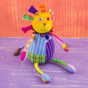 May include: A colorful patchwork lion stuffed animal with a yellow head, blue and purple stripes, and a green and yellow plaid pattern. The lion has a single black eye and a pink nose.