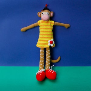 May include: A brown fabric monkey doll with a pink face, wearing a yellow and orange striped dress with a white flower applique. The doll has long legs with white crisscross lacing patterns and wears bright red shoes with white laces.